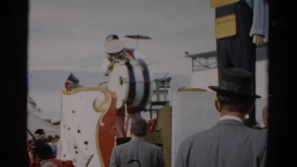 Man in black fedora hat stands back-to-camera in foreground as a decorated band float with large bass drum fills the frame at a 1957 Catskill, New York fair.