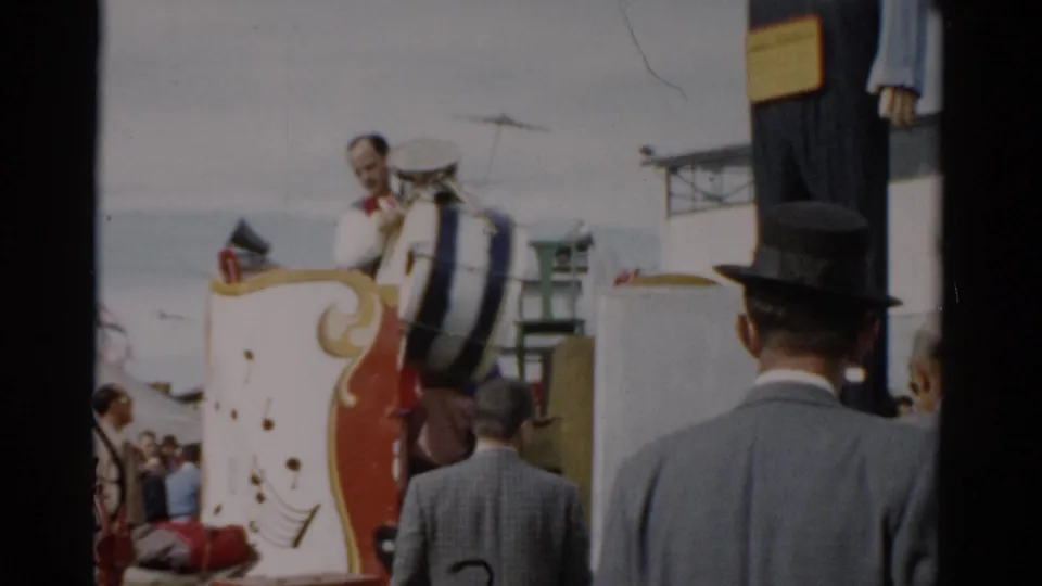 A man in a white shirt visible behind a large bass drum on a red and gold decorated float at what appears to be the 1957 Catskill, New York fair, with a crowd of adults in period attire.