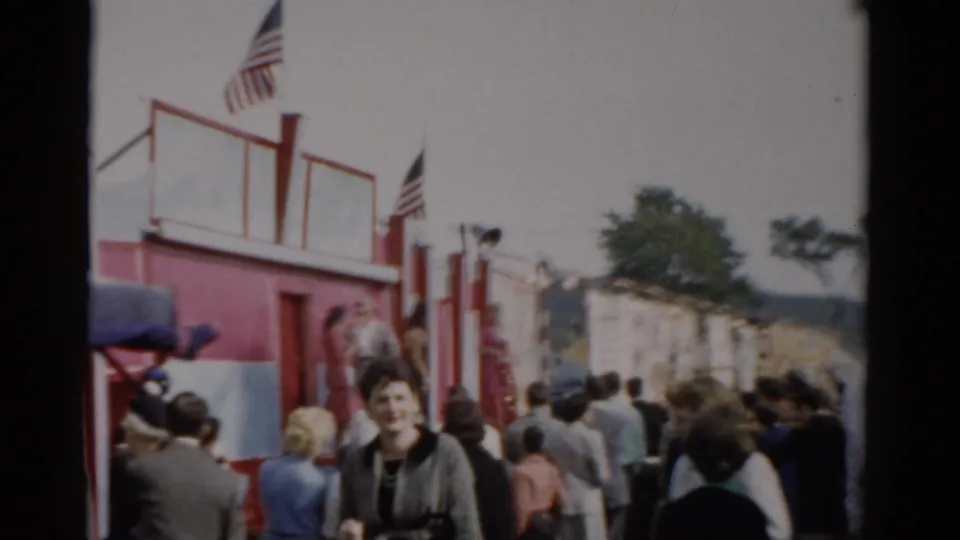 Adults in 1950s dress—including a young woman facing the camera—crowd a fair midway with red-painted structures and American flags, consistent with a 1957 Catskill, New York fair.