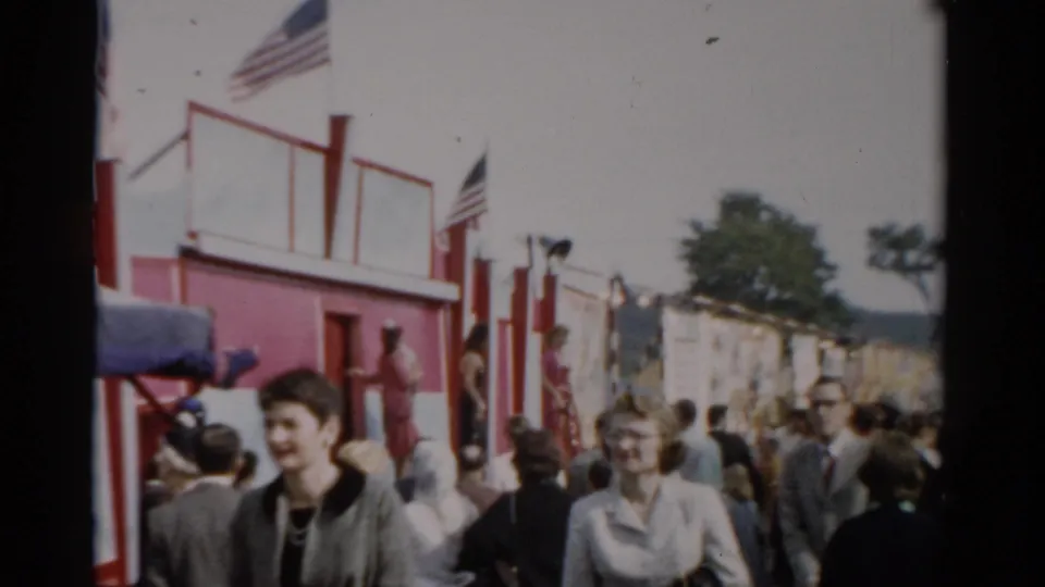 A smiling young woman stands among fair-goers on a midway lined with red structures and American flags at what appears to be the 1957 Catskill, New York fair.