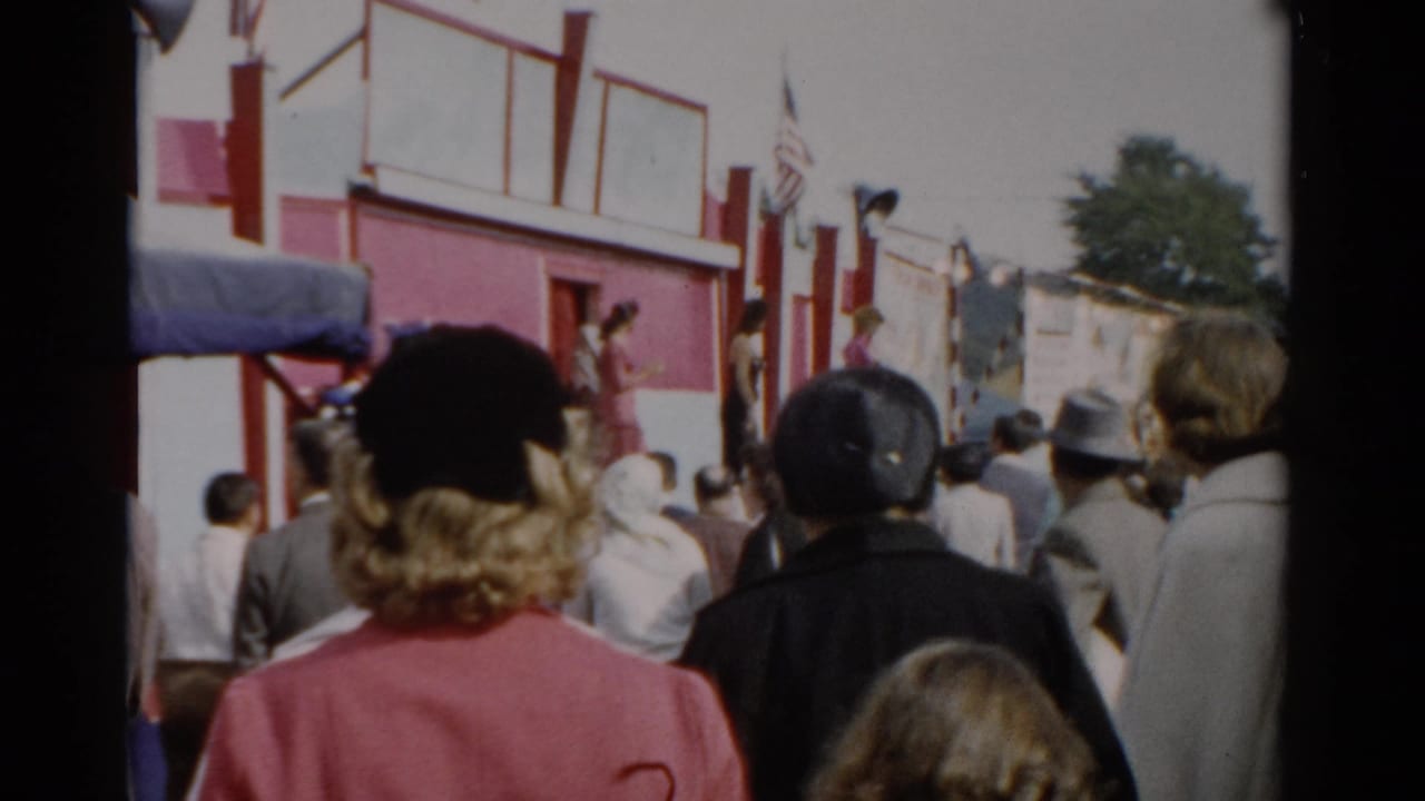 Crowd of fairgoers viewed from behind at what appears to be a 1957 upstate New York fair; red and white building with American flags visible.