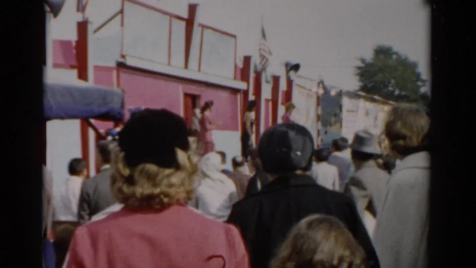 Crowd of fairgoers viewed from behind at what appears to be a 1957 upstate New York fair; red and white building with American flags visible.