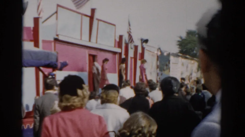 Dense crowd of fairgoers from behind at what appears to be a 1957 upstate New York fair, red and white building with American flags, partial face at right frame edge.