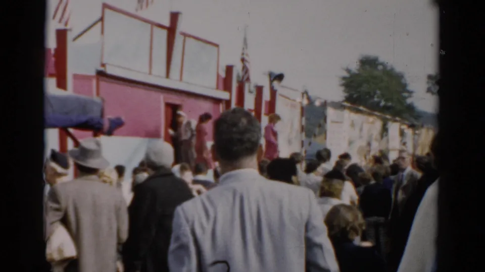 Crowd at what appears to be a 1957 upstate New York fair, red and white building with American flag, people in mid-century dress including man in brimmed hat.