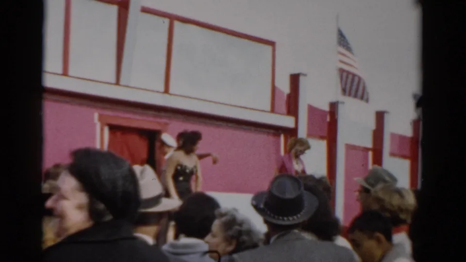 Fairground audience in 1950s attire watches a female entertainer on a pink-and-white stage, American flag overhead; Catskill, New York, c. 1957.