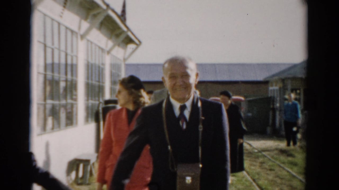 Man in dark suit and tie with shoulder bag walks toward camera; woman in red coat follows, with other visitors and a large building behind them.