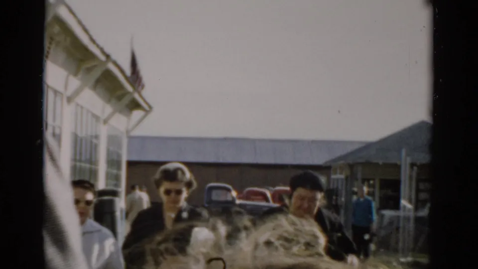 Visitor in sunglasses appears near a large animal at what may be the Catskill Game Farm, c. 1957; late-1950s cars and an American flag visible in background.