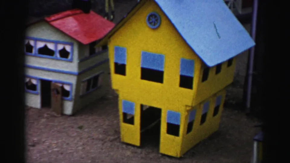Yellow wooden miniature building with blue-trimmed windows and blue dormer, with a red-roofed blue structure behind; part of a 1950s Catskill, New York tourist display on bark mulch.
