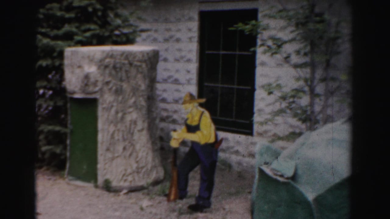 Man in yellow shirt and wide-brimmed hat stands near a stone building with green door; large green tortoise shell structure partially visible at right.