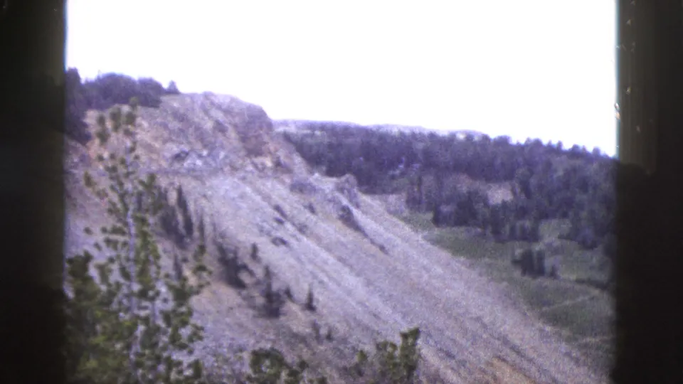 8mm frame circa 1958 showing a wide rocky hillside with a dense conifer treeline along the upper ridge under a bright, washed-out sky, consistent with Yellowstone, Wyoming.