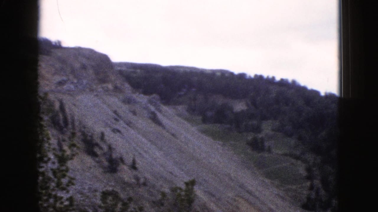 8mm home-movie frame from 1958 Yellowstone area showing a steep eroded hillside, sparse conifers, and pale overcast sky.