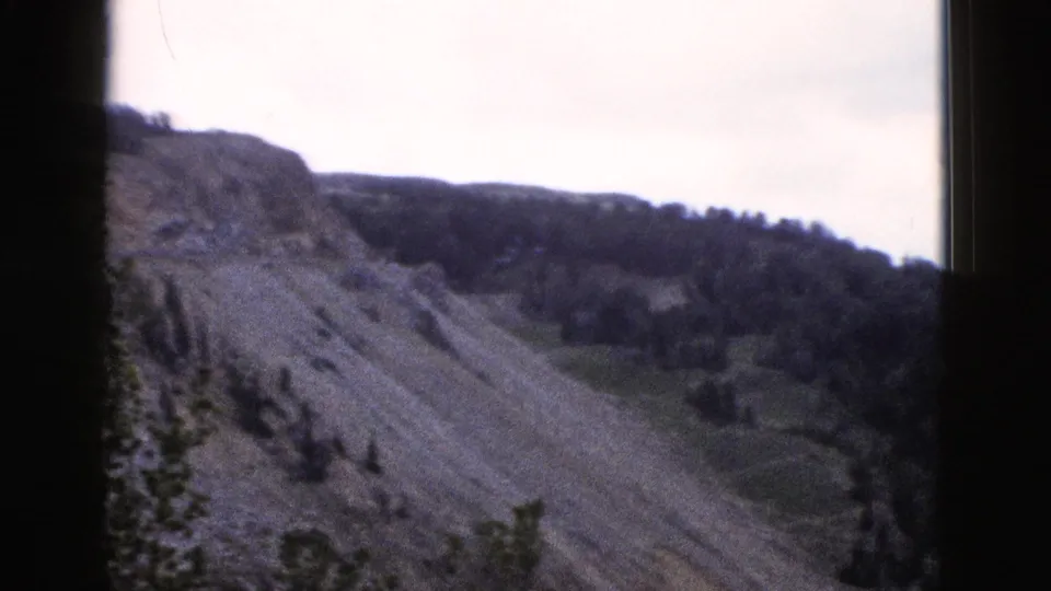 8mm home-movie frame from 1958 Yellowstone area showing a steep eroded hillside, sparse conifers, and pale overcast sky.