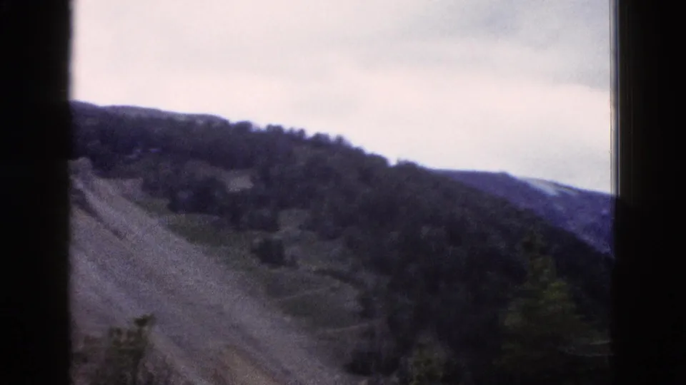8mm home-movie frame from 1958 showing a rocky slope with conifers along the ridge and a distant mountain range visible to the right.