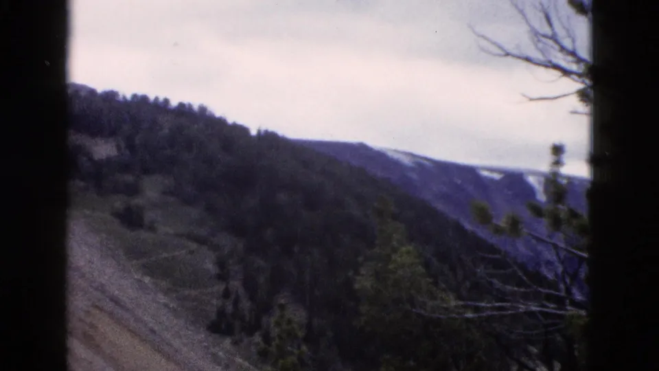 8mm home-movie frame from 1958 Yellowstone area showing a wide mountain valley with snow-covered distant peaks under overcast sky.