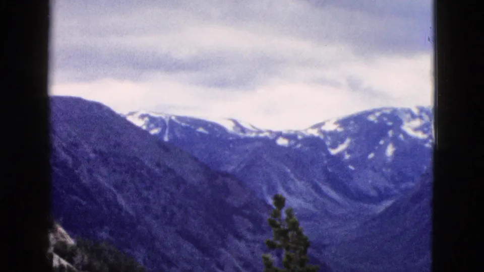 8mm home-movie frame from 1958 Yellowstone showing snow-patched mountain peaks with a cool blue color cast and heavy cloud cover.