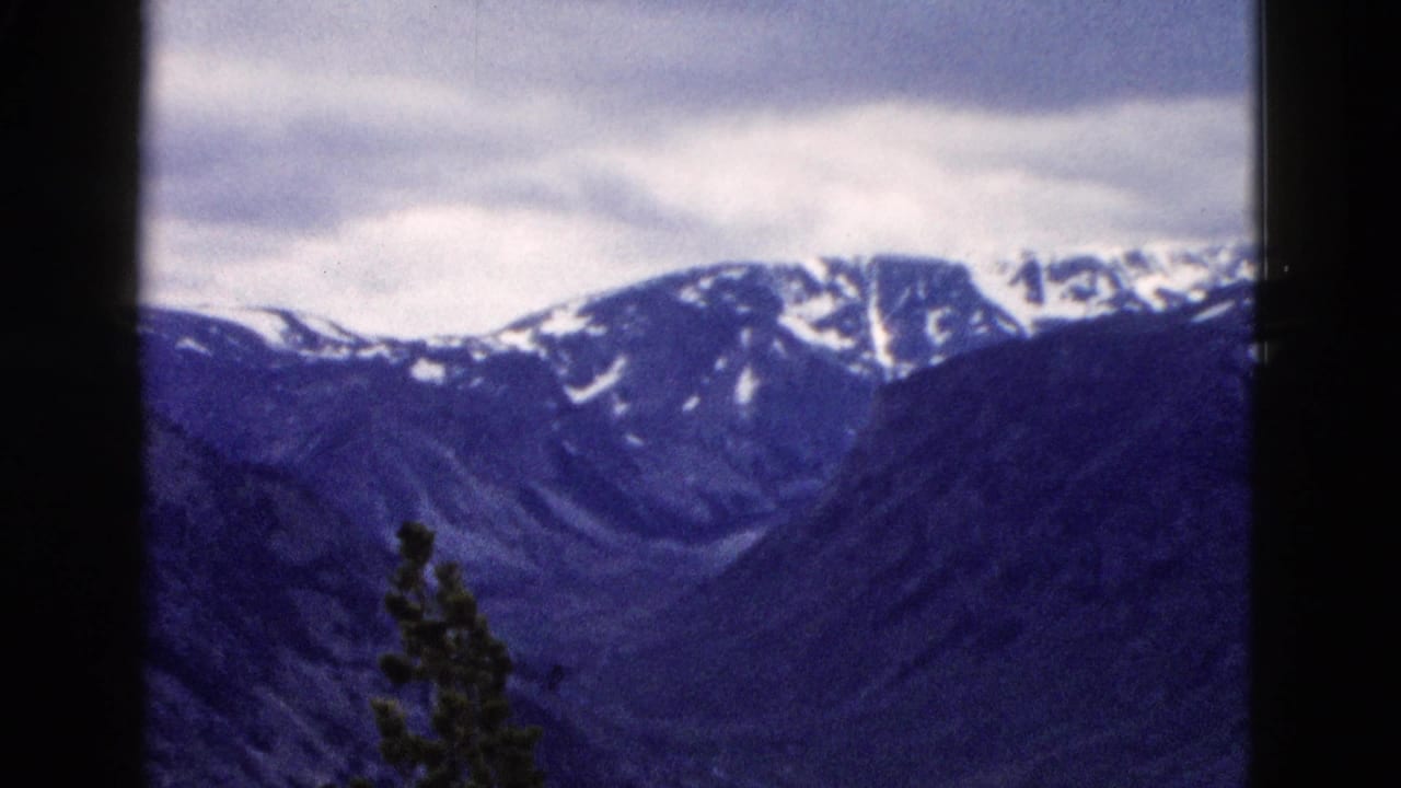 8mm home movie frame circa 1958 showing a snow-patched mountain ridgeline, possibly Yellowstone area Wyoming, under an overcast sky with blue-shifted color.