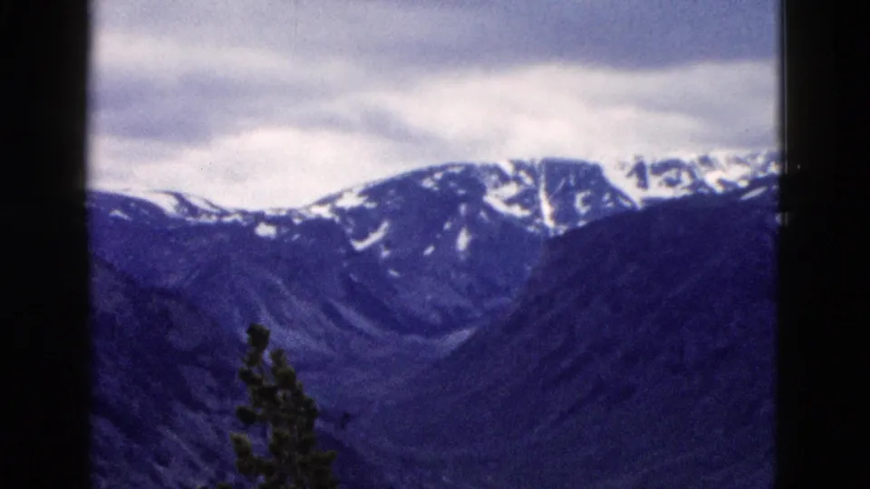 8mm home movie frame circa 1958 showing a snow-patched mountain ridgeline, possibly Yellowstone area Wyoming, under an overcast sky with blue-shifted color.