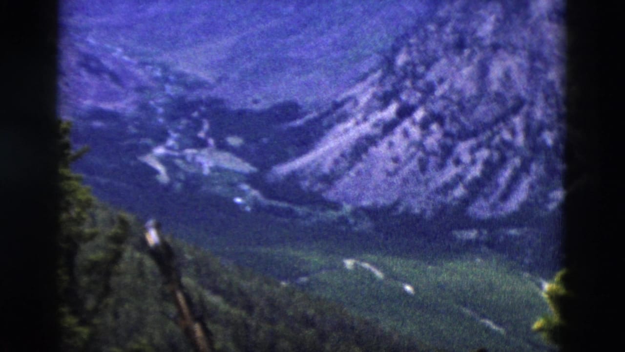 8mm frame circa 1958 of a rugged mountain range with snow remnants on upper slopes and pine trees, viewed from a high-elevation overlook.