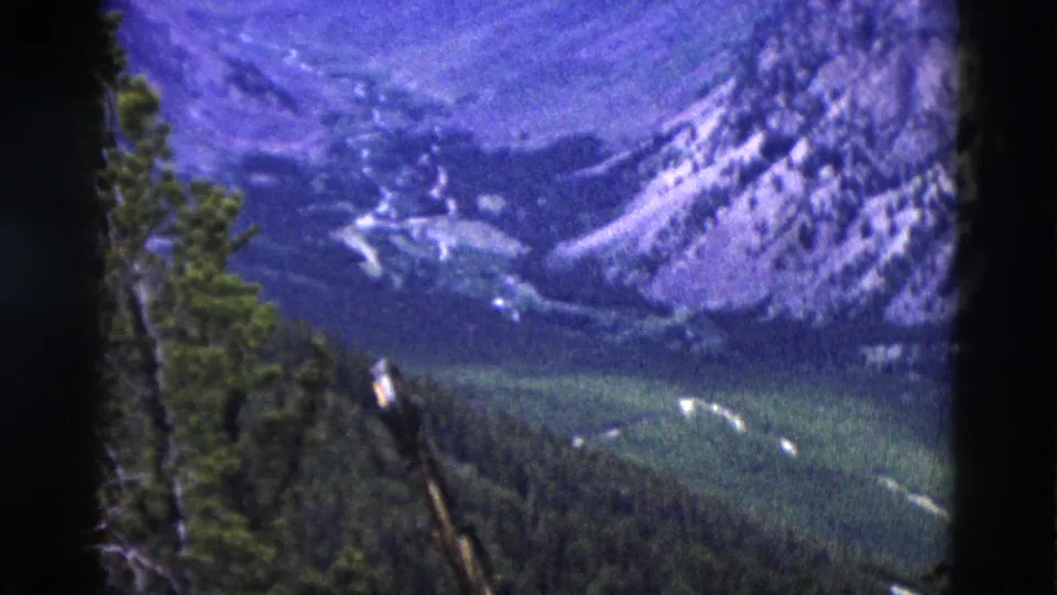Sweeping 8mm view of a rocky mountain range with purple-blue aged-emulsion cast and evergreen-forested valley below, Yellowstone area, Wyoming, 1958.