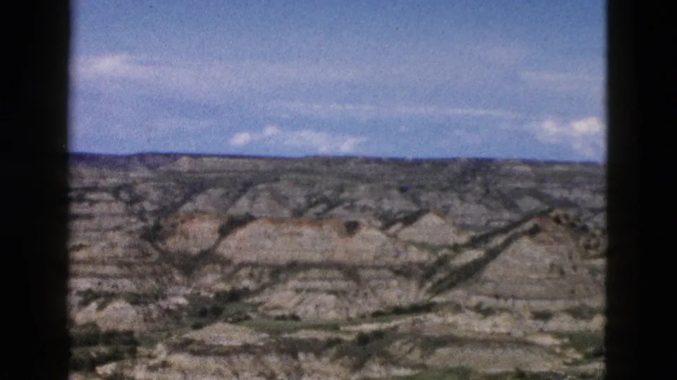 Layered grey and tan eroded buttes in Wyoming, ca. 1958, shot through a car window on 8mm color film with scattered clouds overhead