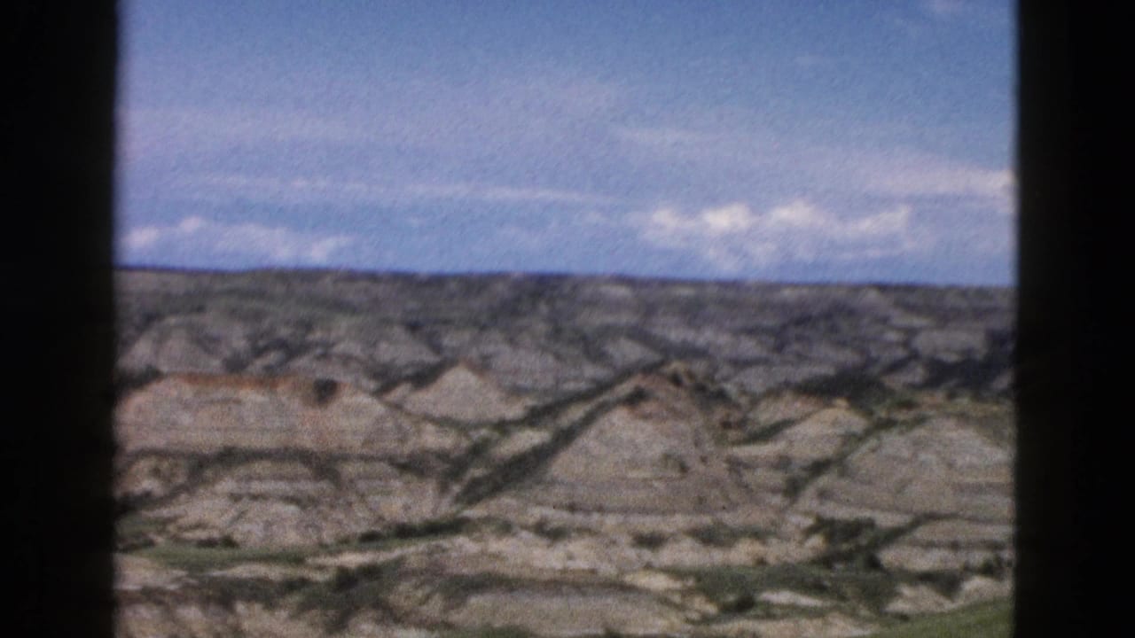Eroded badlands-style buttes in Wyoming, ca. 1958, car-window view showing layered rock formations under partly cloudy blue sky