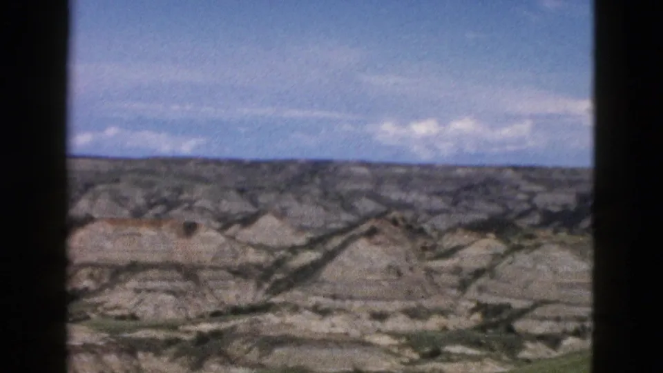 Eroded badlands-style buttes in Wyoming, ca. 1958, car-window view showing layered rock formations under partly cloudy blue sky