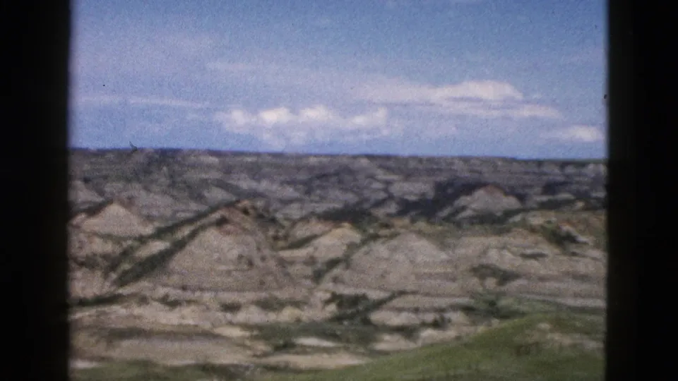 Broader panorama of eroded Wyoming buttes, ca. 1958, showing green low vegetation at the base of grey-tan formations, shot through car window on 8mm film