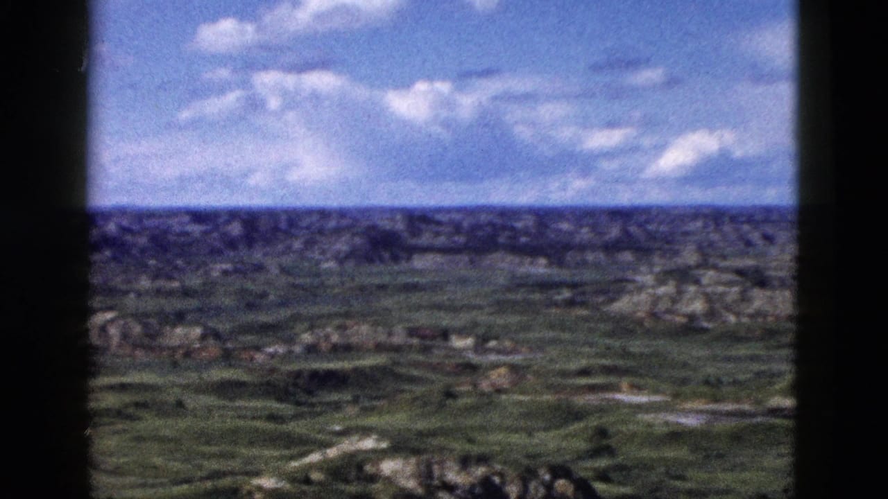 Panoramic 8mm film view of eroded geological formations and green valleys under a partly cloudy sky.