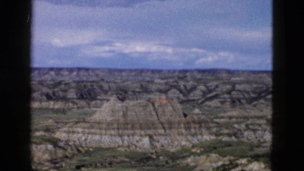 Continued pan across eroded badlands-style terrain with layered buttes and cloudy sky.