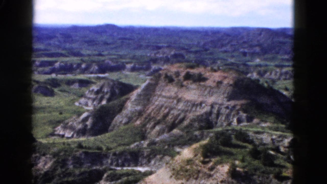 Slow pan continues across a prominent layered rock face with a green valley below and distant mountains.