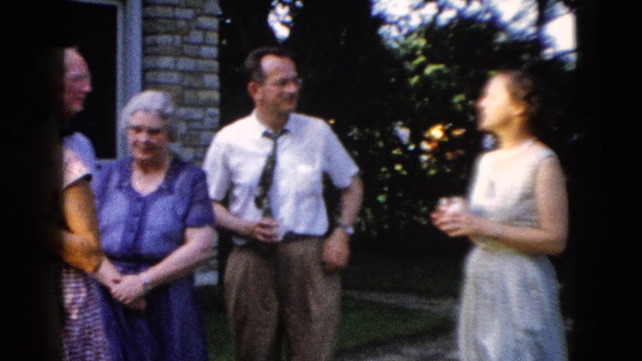 Three adults near a brick wall — man in shirt and tie, woman in light dress, older woman in blue dress — in outdoor conversation.