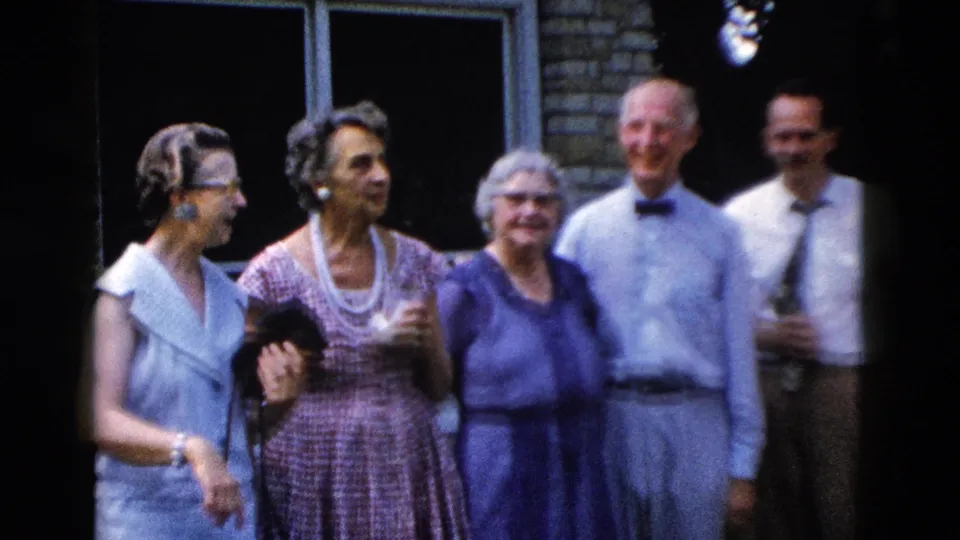 Five adults in late-1950s social attire laughing outdoors; woman in blue dress at left, man with bow tie at center-right; 8mm home movie, Wyoming, circa 1958.