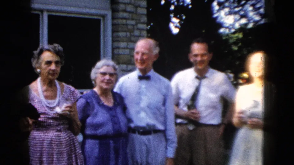 Four adults in 1950s dress attire outside a masonry building; a fifth figure enters from the right, partially overexposed by film edge flare; 8mm home movie circa 1958.