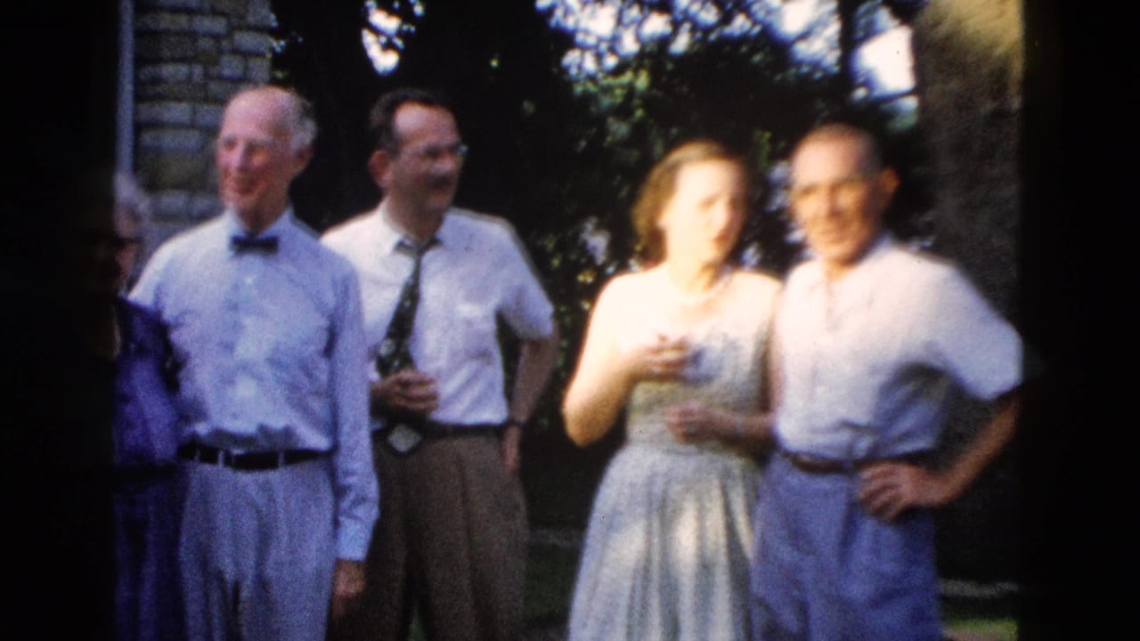 Four adults posed outdoors; men in dress shirts and ties, a woman in a gray-blue full-skirt dress, with a brick building and large tree visible behind.