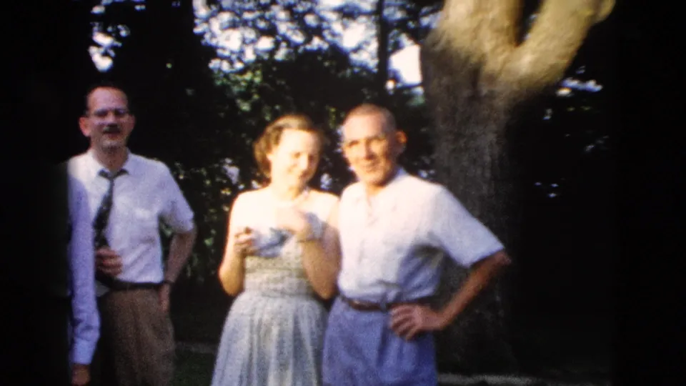 Three adults in late-1950s attire pose outdoors; large deciduous tree trunk and brick building edge visible in background; attributed to Wyoming, 1958.