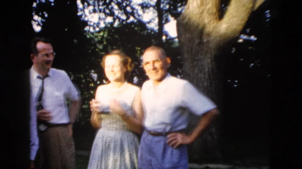 Three adults in late-1950s dress pose together outdoors with a large tree trunk and partially visible brick structure; attributed to Wyoming, 1958.