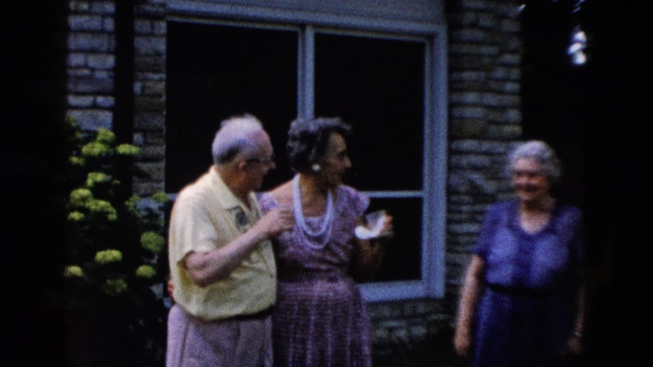 Older man in a yellow shirt and woman in a patterned dress with pearl necklace talk outdoors beside a stone building, circa 1958.