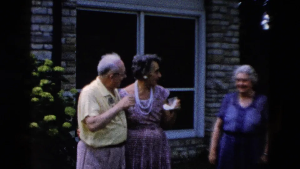 Older man in a yellow shirt and woman in a patterned dress with pearl necklace talk outdoors beside a stone building, circa 1958.