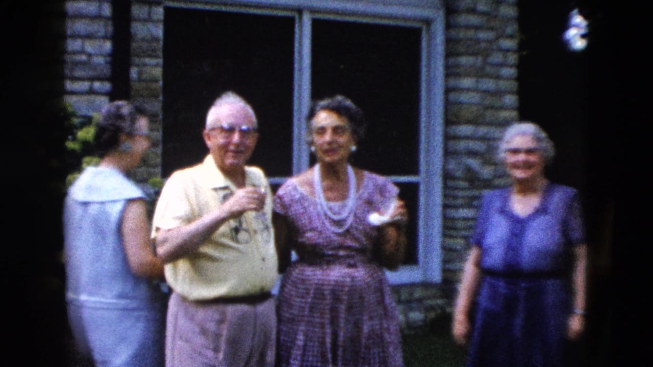 Older man wearing glasses and two women stand beside a stone building; a fourth figure is partially visible at left.