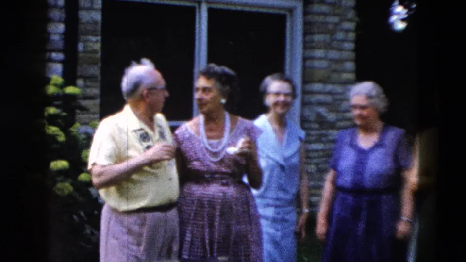 Group of four older adults, including man in yellow shirt and women in patterned and blue dresses, gathered outside a stone building, circa 1958.