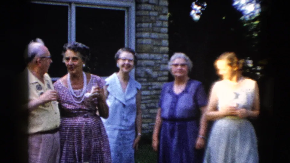Five adults gathered outside a stone building; a younger figure in a light dress appears at far right, group expanding for the camera, circa 1958.