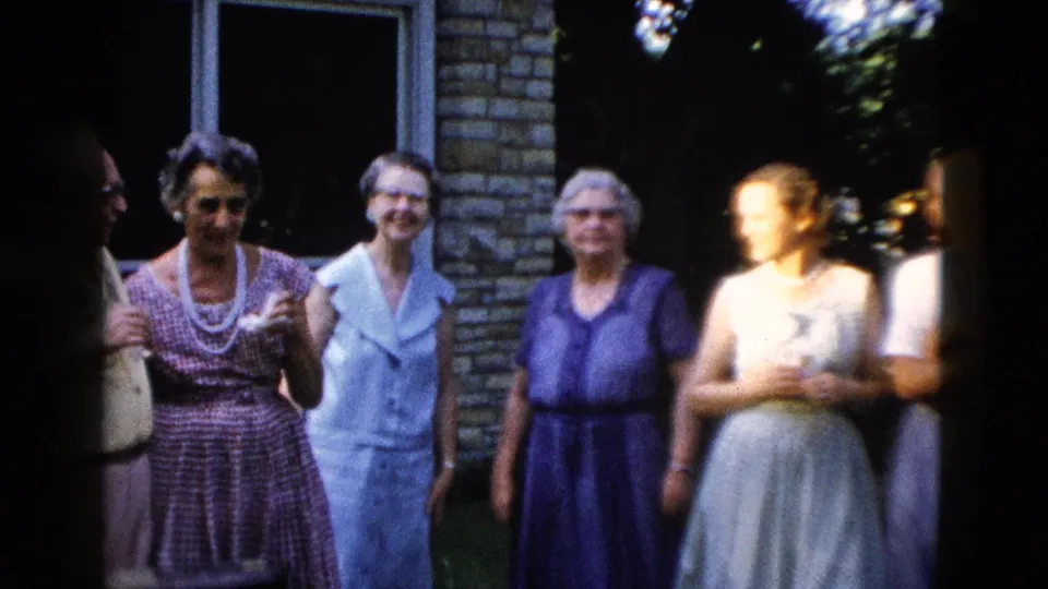 Five or six people, mostly older women in mid-century blue and purple dresses with pearl necklaces, gathered outdoors near a light brick building, circa 1958.