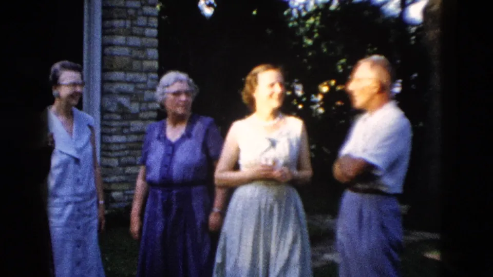 An older man in a light short-sleeved shirt and two older women in blue and purple 1950s dresses stand outdoors near a brick building, circa 1958.