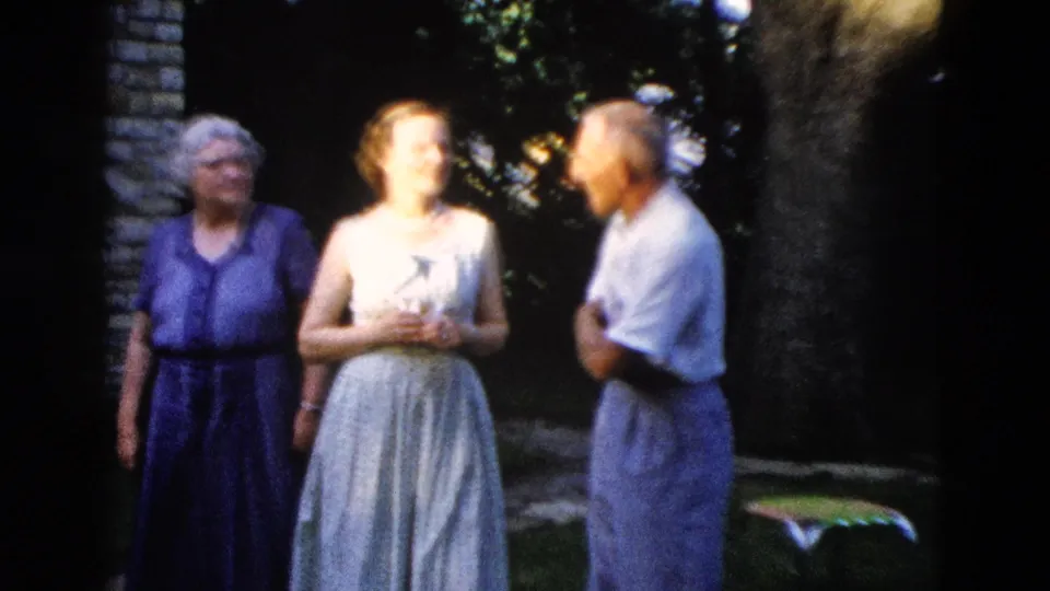 Two older women in blue and purple 1950s dresses and an older man in a light gray shirt engaged in outdoor conversation near a brick building, circa 1958.