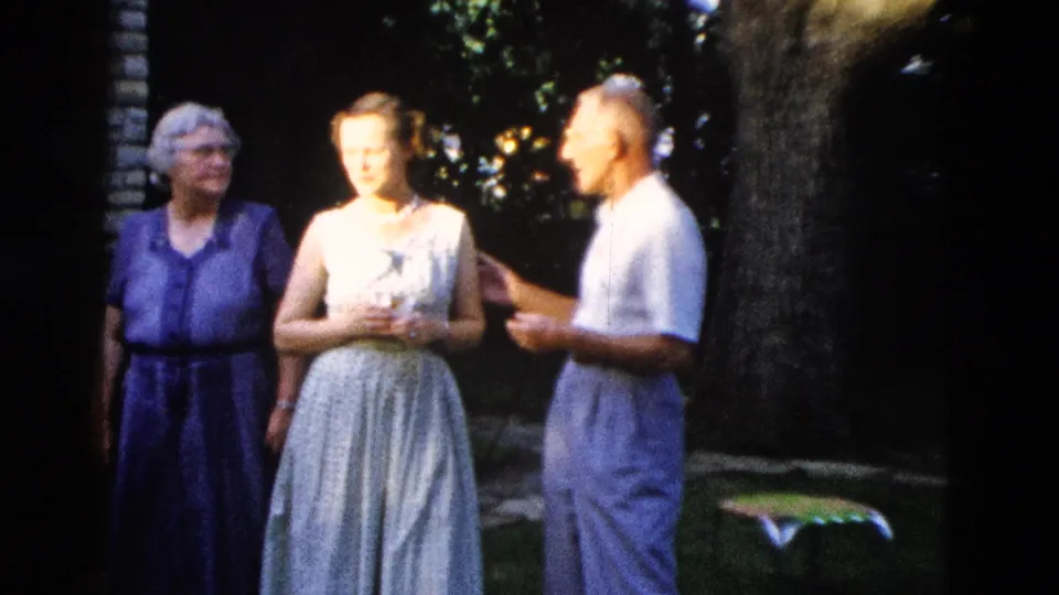 c. 1958 outdoor gathering in Wyoming: elderly woman in purple dress, young woman in light sleeveless dress with pearl necklace, and man in white shirt talking beneath a shade tree.