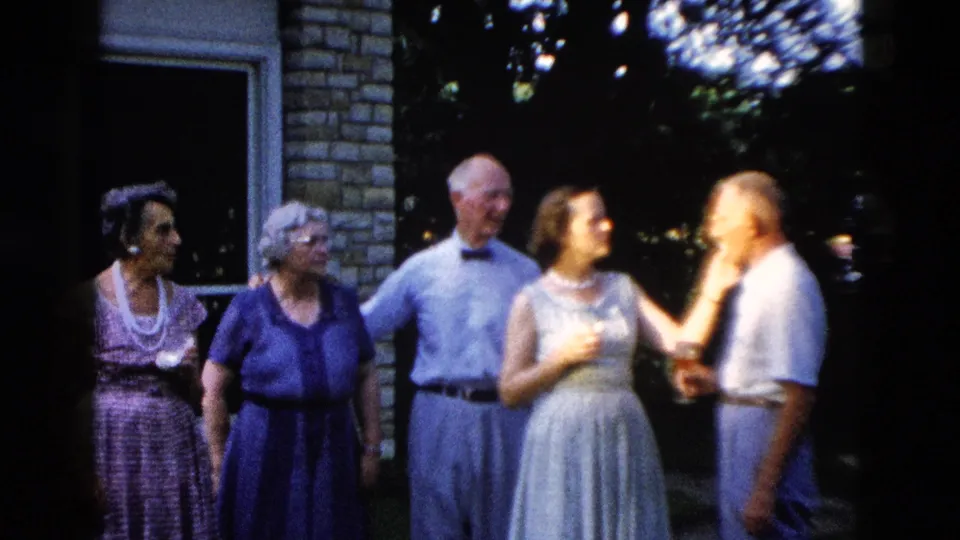 c. 1958 outdoor family gathering in Wyoming: five adults including two elderly women, a bow-tied man, young woman with pearl necklace, and another man stand near a brick building with window and shade tree.