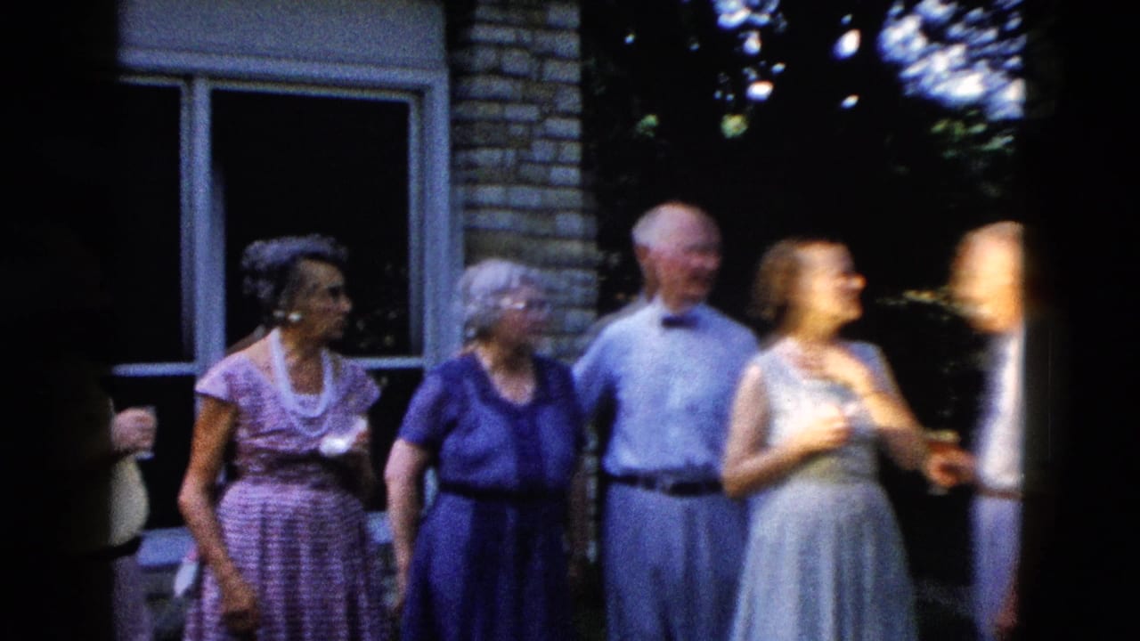 Five older adults standing in front of a brick house with large windows; women in mid-length dresses, men in collared shirts; hydrangea bushes at right.