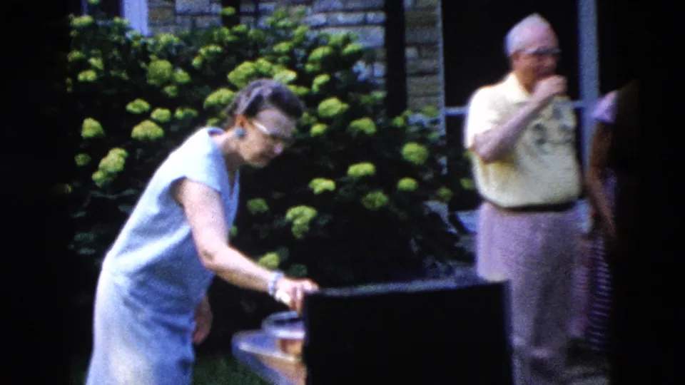 Woman reaching toward what appears to be a cooler or container at a backyard social gathering, c. 1958; man in background drinking, hydrangeas and brick house prominent.