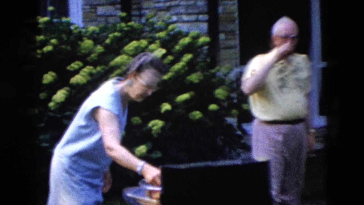 A woman in a light blue dress leans toward what may be a grill or dish while an older man in a yellow shirt stands nearby — 1958 backyard gathering, possibly Wyoming.
