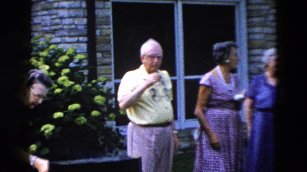 Older man in yellow shirt stands near a brick wall beside a woman in a purple dress; a third figure is partially visible at right.